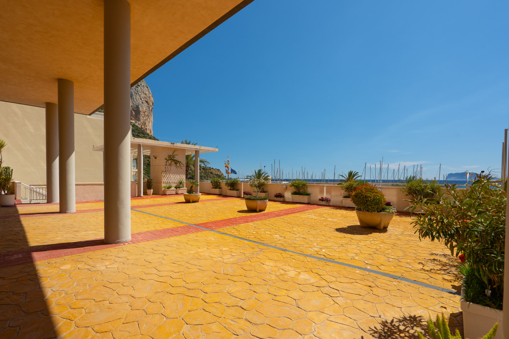 Zona exterior común con pavimento texturizado amarillo y rojo, plantas en macetas, y vistas directas al puerto deportivo con mástiles de barcos y al emblemático Peñón de Ifach.