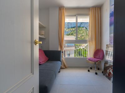 Acceso a luminosa habitación con sofá gris, estantes flotantes y ventana con vistas directas al rocoso Peñón de Ifach. Una silla de oficina rosa complementa el espacio.