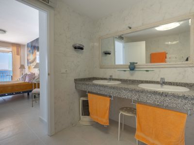 Baño en suite con doble lavabo de granito y azulejos de mármol blanco. A través de la puerta abierta, se vislumbra el dormitorio adyacente con vistas al mar.