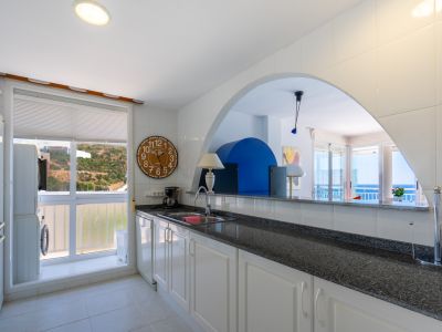 Cocina semiabierta con encimera oscura y armarios blancos, incluyendo una zona de lavandería integrada y un arco que conecta con el salón con vistas al mar.