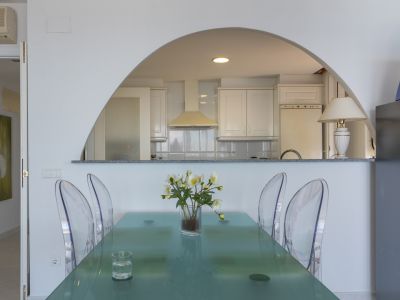 Comedor con mesa de cristal moderna y sillas transparentes, conectado por un arco a la cocina semiabierta con armarios blancos y azulejos.