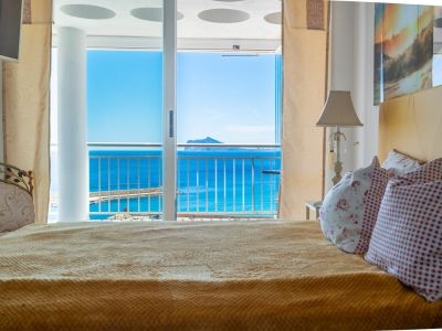 Dormitorio con cama confortable y acceso directo a balcón, ofreciendo vistas panorámicas al mar azul, la marina y una isla lejana.