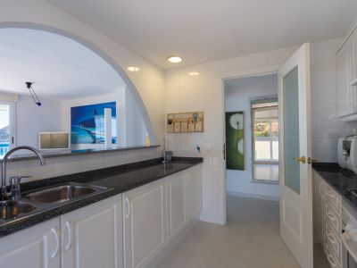 Luminosa cocina con azulejos blancos, encimeras oscuras y doble fregadero, comunicada por un arco con vistas al mar y a la montaña.
