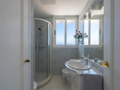 Luminoso baño con cabina de ducha curva de cristal, azulejos de mosaico y tocador blanco con encimera de mármol bajo una ventana que aporta luz natural.