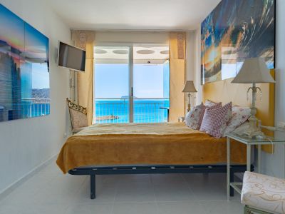 Luminoso dormitorio con cabecero amarillo y acceso directo a terraza mediante puertas correderas de cristal, ofreciendo vistas al mar azul y una masa de tierra distante.