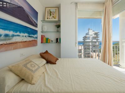 Luminoso dormitorio con cama, arte mural de playa, estanterías de cristal y acceso a balcón con vistas al mar y edificios.