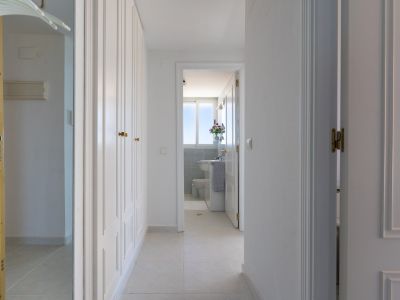 Luminoso pasillo con armarios empotrados blancos y tiradores dorados. El suelo de baldosas conduce a un baño visible a través de una puerta abierta, mostrando una ventana y azulejos de mosaico azul.