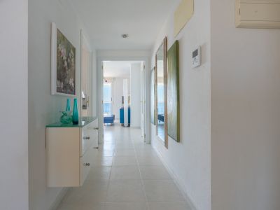 Luminoso pasillo con suelo de baldosas y consola flotante de cristal. Conduce a una sala soleada con vistas al mar, decorado con jarrones y arte mural.