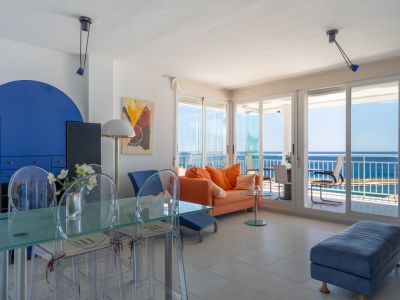 Luminoso salón-comedor con sofá naranja, mesa de cristal y acceso directo a una terraza acristalada con vistas panorámicas al mar.