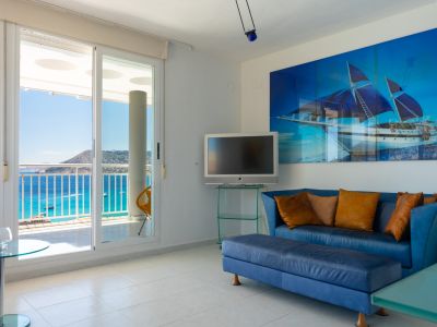 Salón luminoso con sofá azul, obra de arte de velero y puertas correderas que dan a una terraza con vistas panorámicas al mar y la costa.