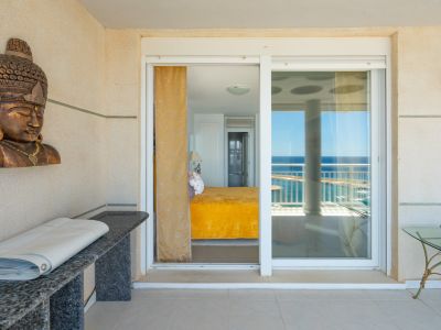Vista desde la terraza a un dormitorio con colcha amarilla brillante, destacando un busto de madera tallada y una consola de granito. Puertas correderas de cristal reflejan el mar.
