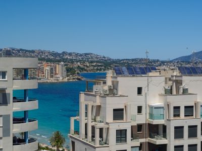 Vista elevada de edificios costeros modernos con balcones sobre el mar turquesa, la costa lejana y montañas bajo un cielo despejado. Paneles solares visibles en los tejados.