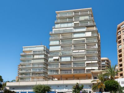 Vista exterior de un moderno edificio residencial de varias plantas con numerosos balcones, muchos cerrados con cortinas de cristal y barandillas blancas, bajo un cielo azul claro. Palmeras en primer plano.