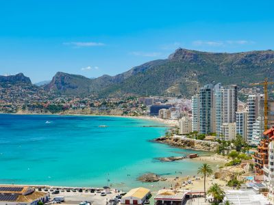 Vista panorámica de la bahía de Calpe con aguas turquesas, playas de arena, modernos edificios costeros y montañas verdes bajo un cielo despejado.