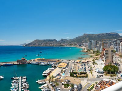 Vista panorámica del animado puerto deportivo de Calpe con numerosos yates, la bahía turquesa, la playa de arena y las montañas al fondo.