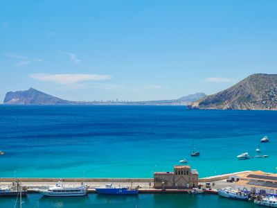 Vista panorámica del puerto deportivo de Calpe con embarcaciones, aguas turquesas y montañas lejanas. El Peñón de Ifach domina a la derecha, con un skyline urbano al fondo.