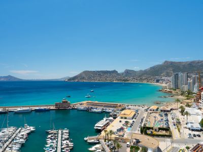 Vista panorámica del puerto deportivo de Calpe con yates, la vibrante bahía de aguas turquesas y una playa de arena junto a edificios costeros y montañas.