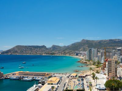 Vista panorámica del puerto deportivo de Calpe, la bahía turquesa y la Playa de La Fossa, enmarcada por montañas lejanas bajo un cielo azul claro.