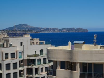 Vistas panorámicas al mar desde una planta alta, con arquitectura moderna adyacente, paneles solares y una costa montañosa urbanizada a lo lejos.