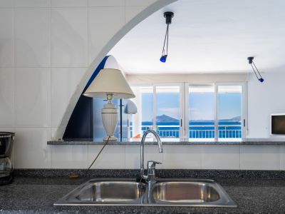 Zona de cocina con fregadero doble de acero inoxidable y encimera moteada, enmarcando vistas al mar y montañas lejanas a través de un arco y puertas correderas de cristal.