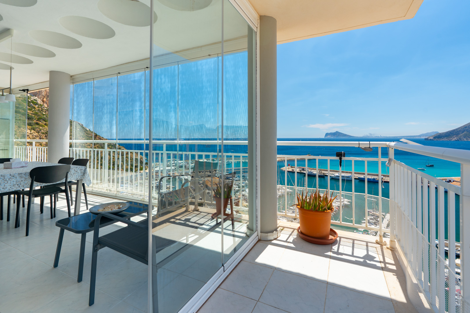 Amplia terraza con cortinas de cristal que encierran un comedor, abriéndose a un balcón. Ofrece vistas panorámicas al puerto deportivo de Calpe, el mar azul y la costa lejana bajo un cielo despejado.