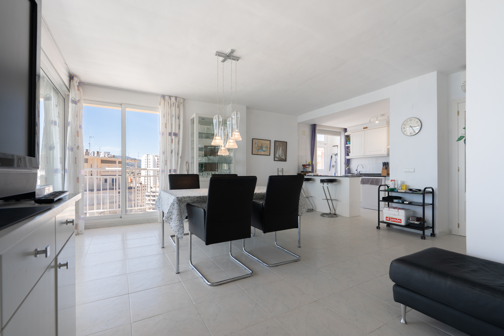 Amplio salón-comedor de planta abierta con mesa moderna, cocina integrada con barra americana y grandes ventanales que ofrecen vistas urbanas.