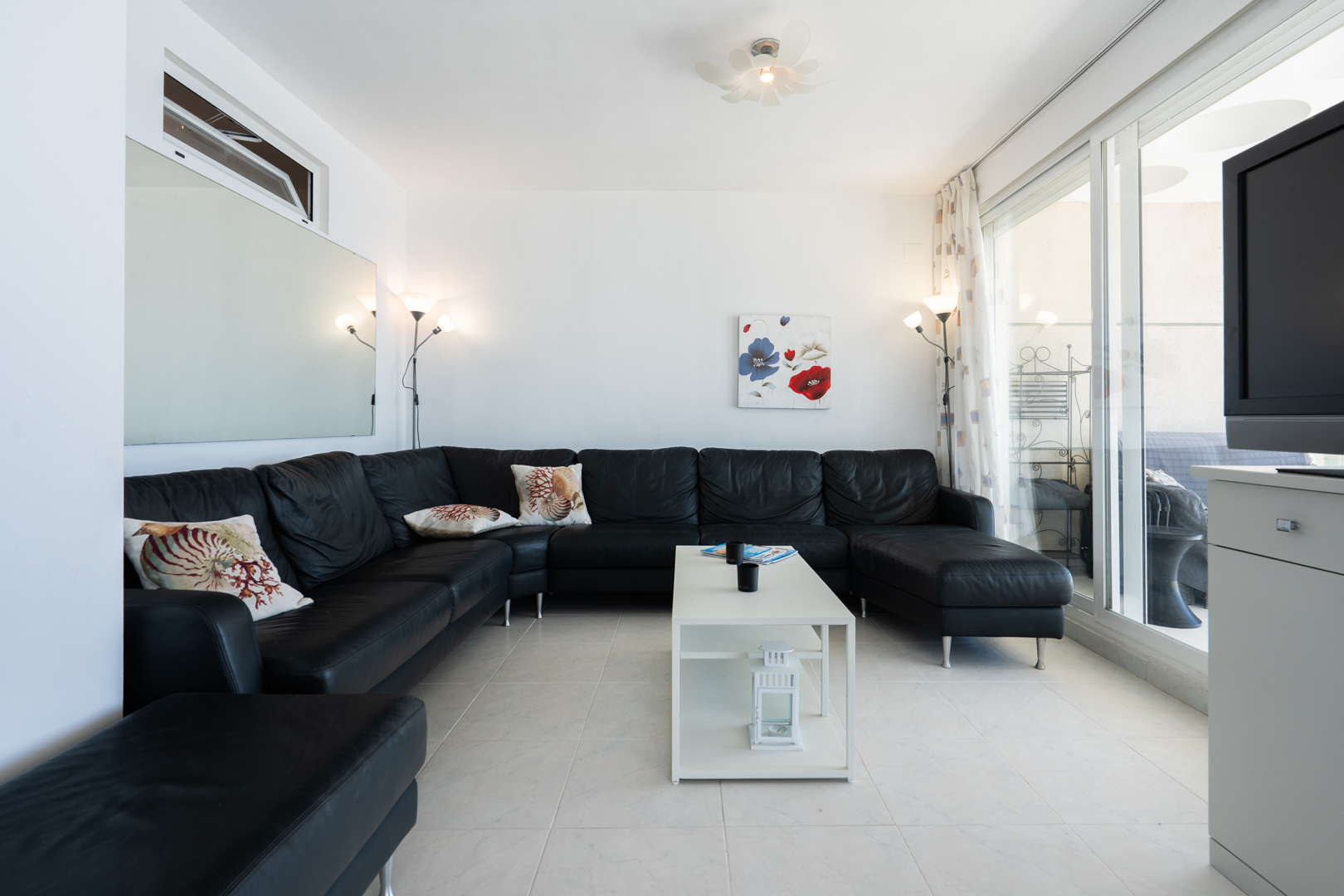 Amplio salón con sofá esquinero de cuero negro, suelo de baldosas blancas y gran espejo. Las puertas correderas de cristal conectan con una terraza cubierta, aportando luz natural.
