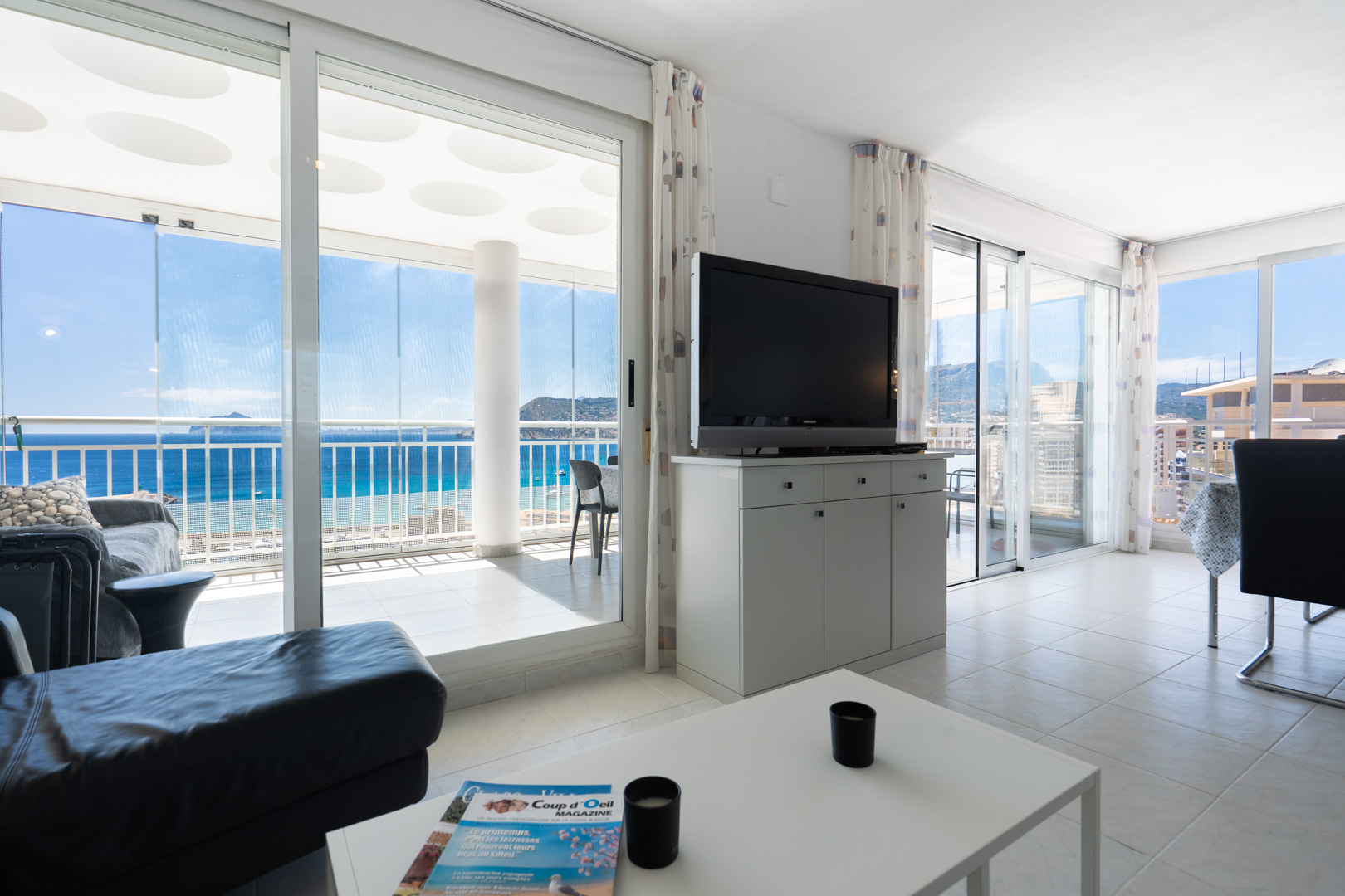 Amplio salón con sofá oscuro y televisor, conectado a una terraza acristalada que ofrece vistas panorámicas al mar, la costa y el horizonte. El suelo es de baldosas claras.