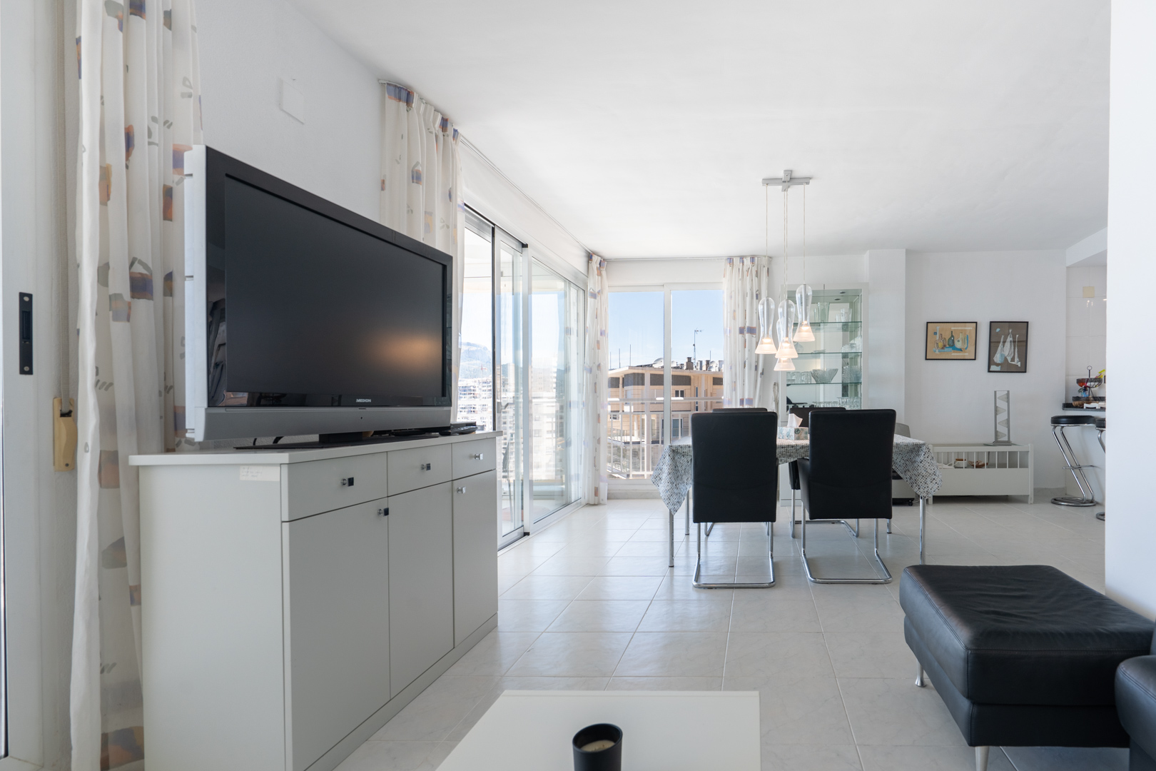 Luminoso salón-comedor con suelos de baldosas blancas, televisor sobre un mueble gris y mesa de comedor con lámparas colgantes modernas. Puertas correderas de cristal dan a un balcón con vistas urbanas.
