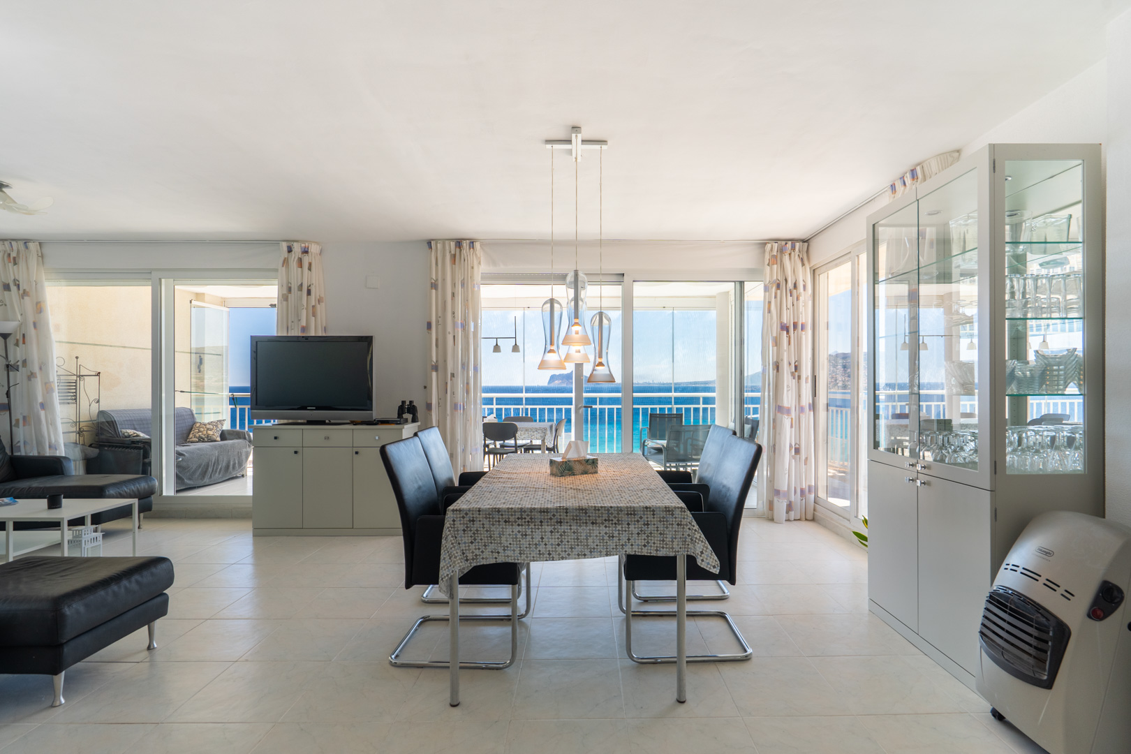 Luminoso salón-comedor diáfano con suelo de baldosas, moderna iluminación colgante y amplias puertas correderas de cristal hacia la terraza con vistas panorámicas al mar y al Peñón de Ifach.