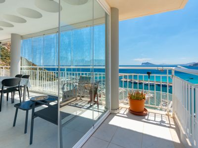 Amplia terraza con cortinas de cristal que encierran un comedor, abriéndose a un balcón. Ofrece vistas panorámicas al puerto deportivo de Calpe, el mar azul y la costa lejana bajo un cielo despejado.