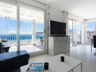 Amplio salón con sofá oscuro y televisor, conectado a una terraza acristalada que ofrece vistas panorámicas al mar, la costa y el horizonte. El suelo es de baldosas claras.