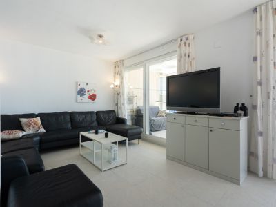 Salón luminoso con sofá seccional de cuero negro, mesa de centro blanca y TV sobre mueble gris. Puertas correderas dan a una terraza cubierta.