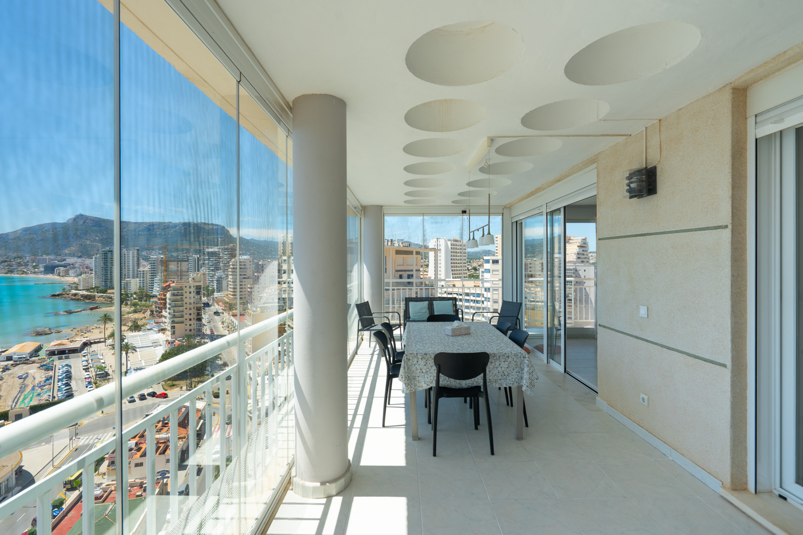Amplia terraza acristalada con comedor y vistas panorámicas al mar, puerto y montañas de Calpe. Destacan las cortinas de cristal, el techo con detalles circulares y el suelo de gres.