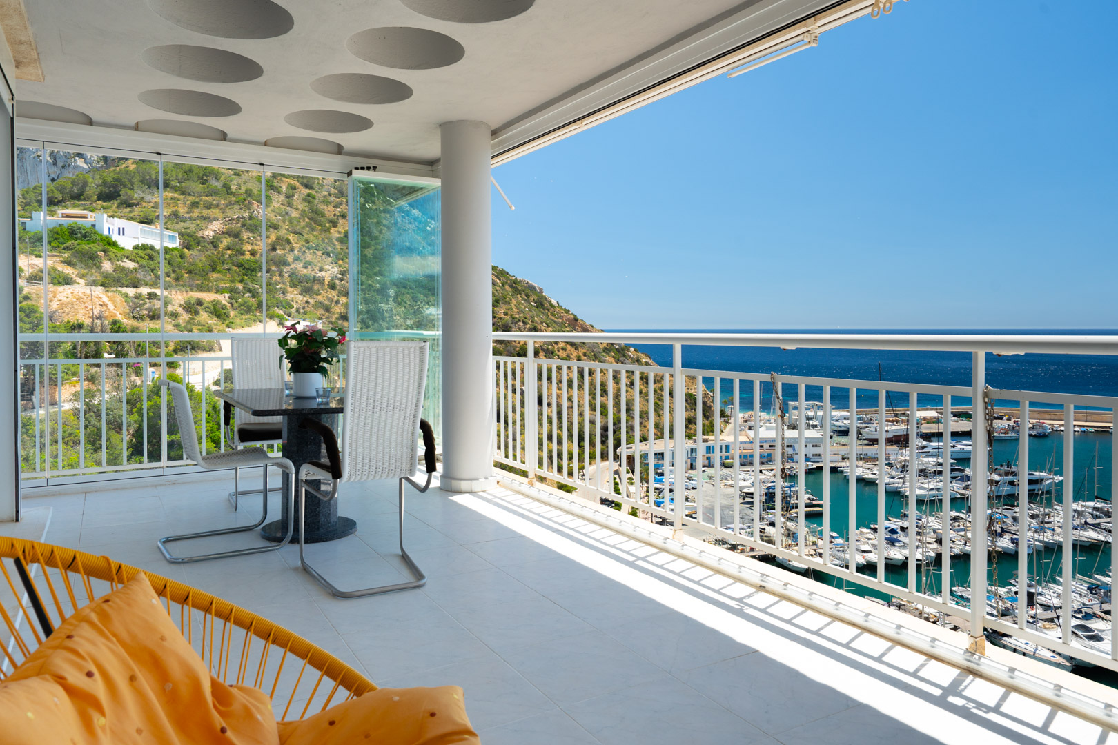 Amplia terraza con suelo de baldosas claras y cortinas de cristal que enmarcan una ladera verde. Vistas abiertas al puerto deportivo y al mar azul, con conjunto de comedor y barandilla blanca.