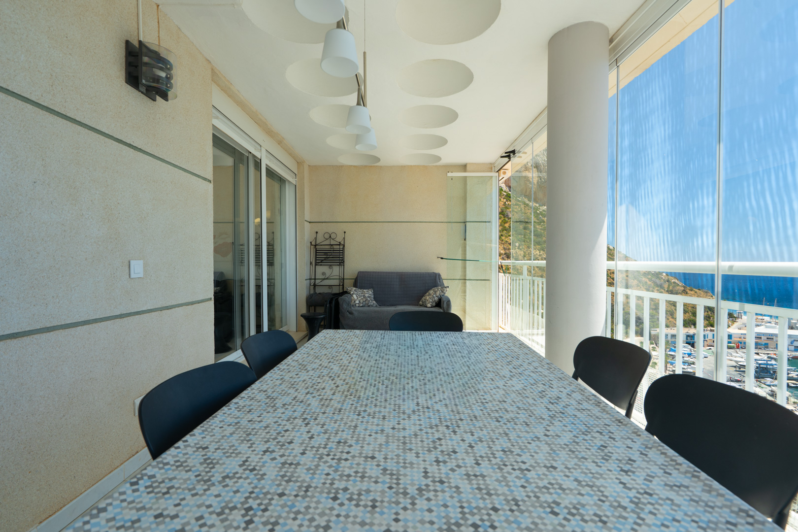 Amplia terraza cubierta con mesa de comedor de mosaico, asientos y cortinas de cristal con vistas panorámicas al puerto, el mar y las montañas.