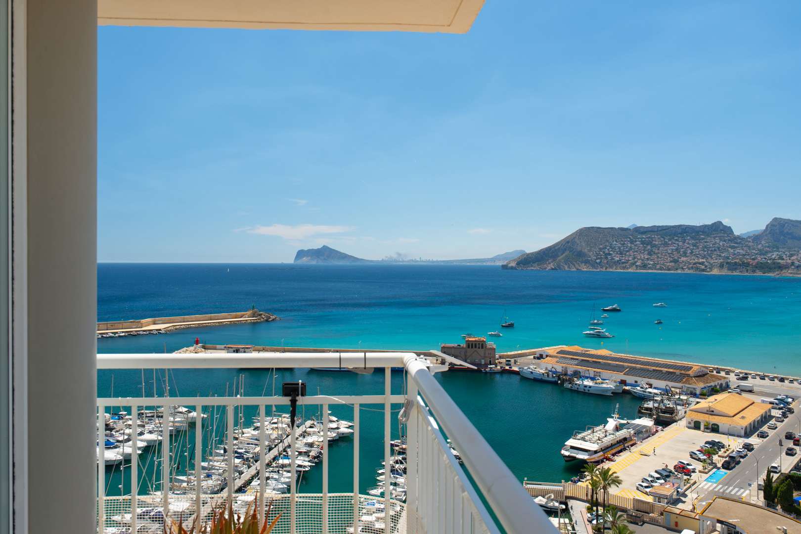 Amplias vistas al mar y al puerto deportivo desde el balcón, con numerosos barcos, la bahía turquesa y montañas lejanas bajo un cielo azul claro.
