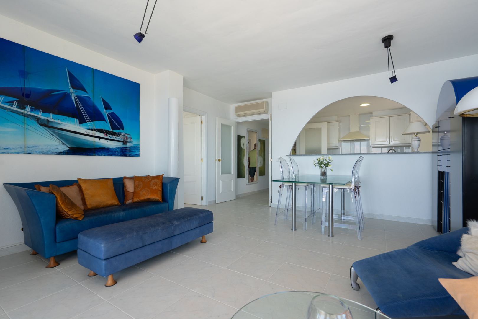 Amplio salón-comedor con sofá azul, mesa de cristal y sillas transparentes, complementado por un gran cuadro de velero. Una arcada conecta con la moderna cocina semiabierta.
