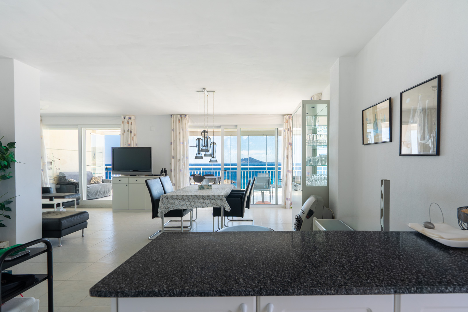 Amplio salón-comedor de planta abierta con lámpara colgante moderna sobre la mesa, suelo de baldosas claras y acceso directo a una terraza con vistas panorámicas al mar a través de puertas correderas de cristal.