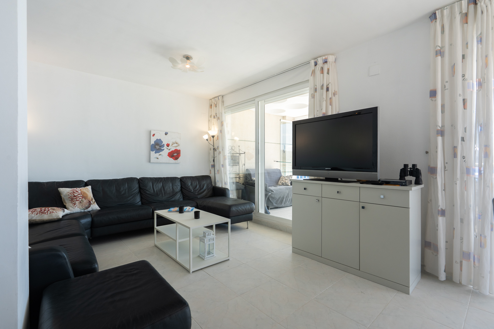 Amplio salón con sofá seccional de cuero negro, mesa de centro blanca y suelo de baldosas. Puertas correderas de cristal dan a una terraza, junto a un mueble con TV y cortinas estampadas.