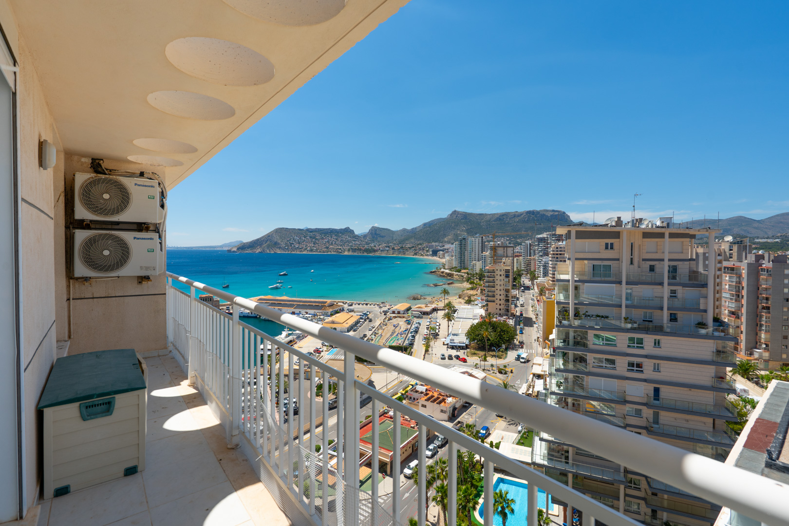 Balcón con vistas panorámicas al puerto deportivo de Calpe, la bahía turquesa y la costa urbana, enmarcado por una barandilla blanca y unidades exteriores de aire acondicionado.