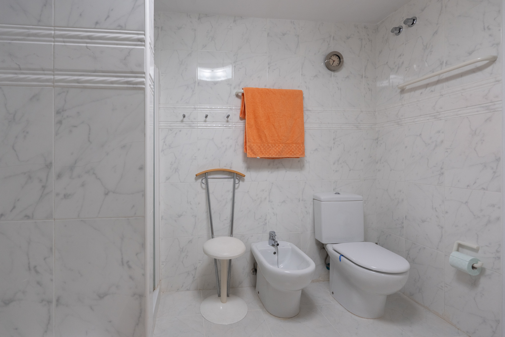 Baño con azulejos efecto mármol blanco, equipado con inodoro, bidé y una práctica zona de ducha con barra de apoyo. Incluye un reloj de pared y perchas para toallas.