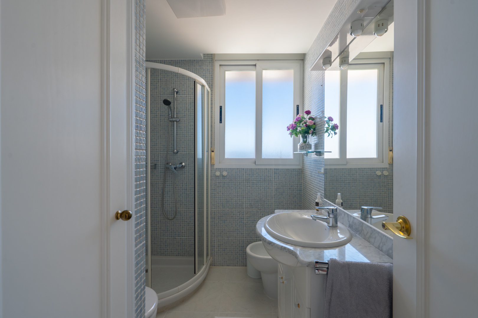 Baño con ducha de esquina, lavabo sobre encimera de mármol y azulejos de mosaico azul claro. Ventana que aporta luz natural y espejo con focos.