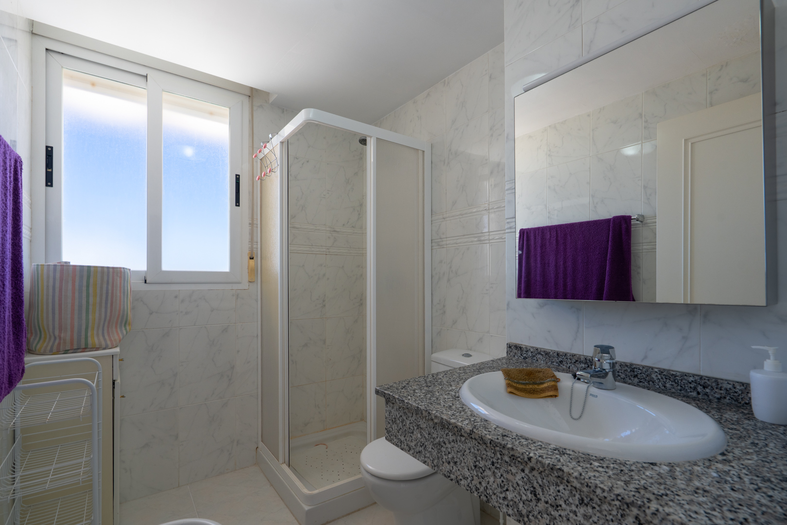 Baño funcional con azulejos efecto mármol blanco, encimera de granito gris y lavabo ovalado. Incluye cabina de ducha de esquina y ventana con luz natural.