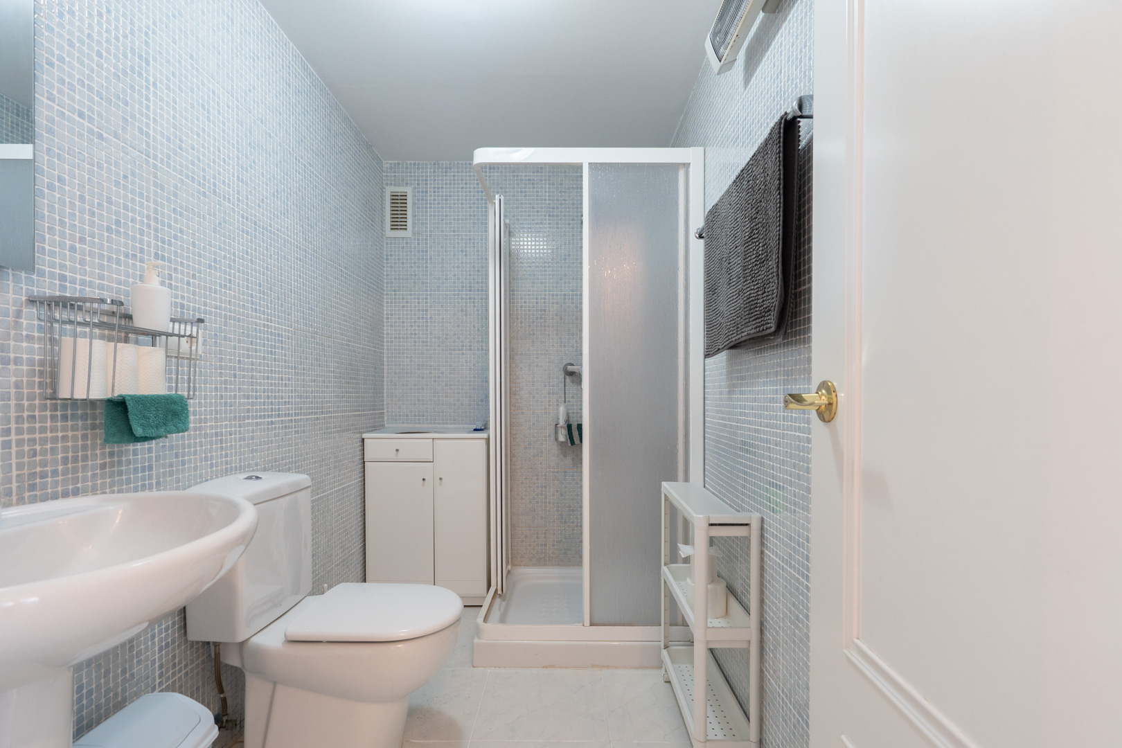 Baño funcional con paredes de mosaico azul claro, equipado con inodoro, lavabo de pedestal y cabina de ducha. Incluye un pequeño mueble de almacenamiento y estanterías auxiliares.