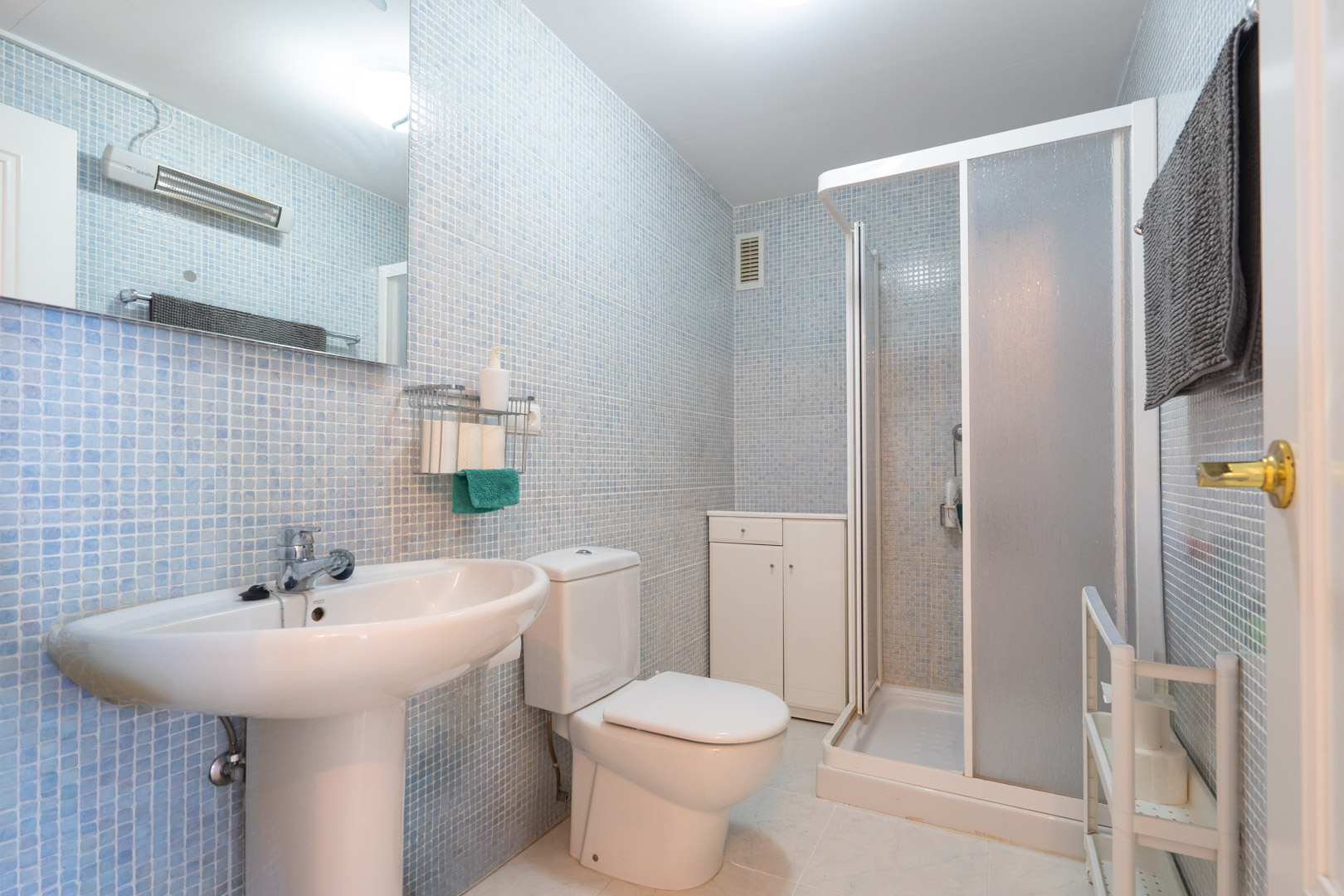 Baño funcional con paredes de mosaico azul claro, lavabo de pedestal, inodoro, ducha con mampara de cristal esmerilado y mueble auxiliar blanco para almacenamiento.