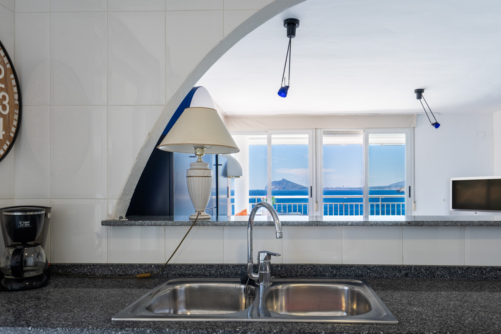 Cocina con fregadero doble y encimera oscura, conectada a un luminoso salón a través de un arco, ofreciendo impresionantes vistas al mar y la costa desde la terraza.