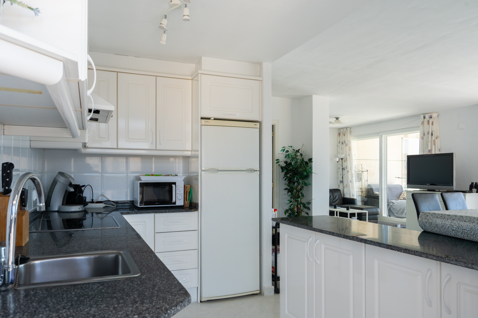 Cocina equipada con armarios blancos panelados, encimeras de granito oscuro moteado y fregadero de acero inoxidable, abierta al salón mediante una barra americana y con luz natural de la terraza.
