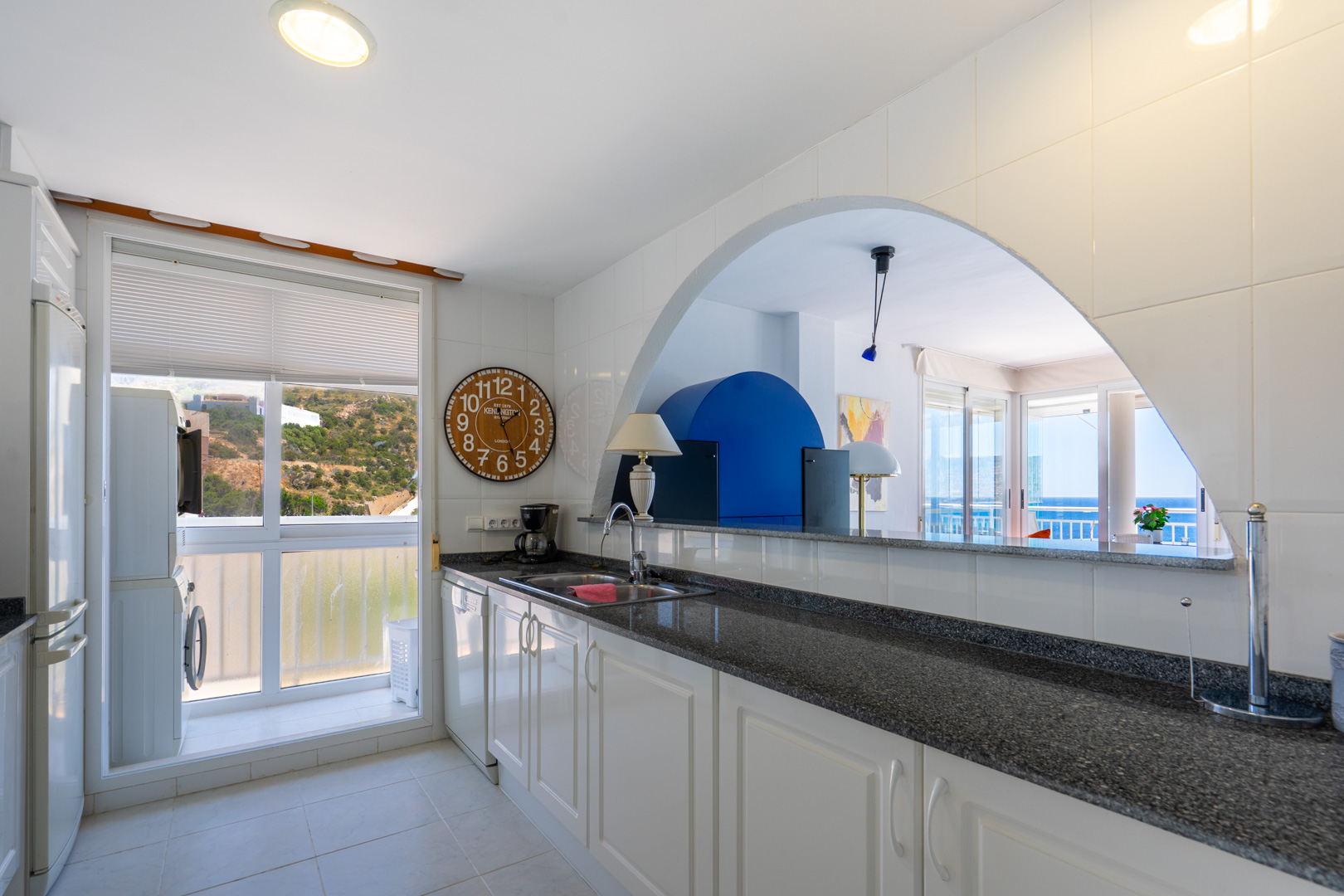 Cocina funcional con armarios blancos, encimera de granito oscuro y zona de lavandería junto a una ventana con vistas a la ladera. Un arco conecta con el salón y vistas al mar.