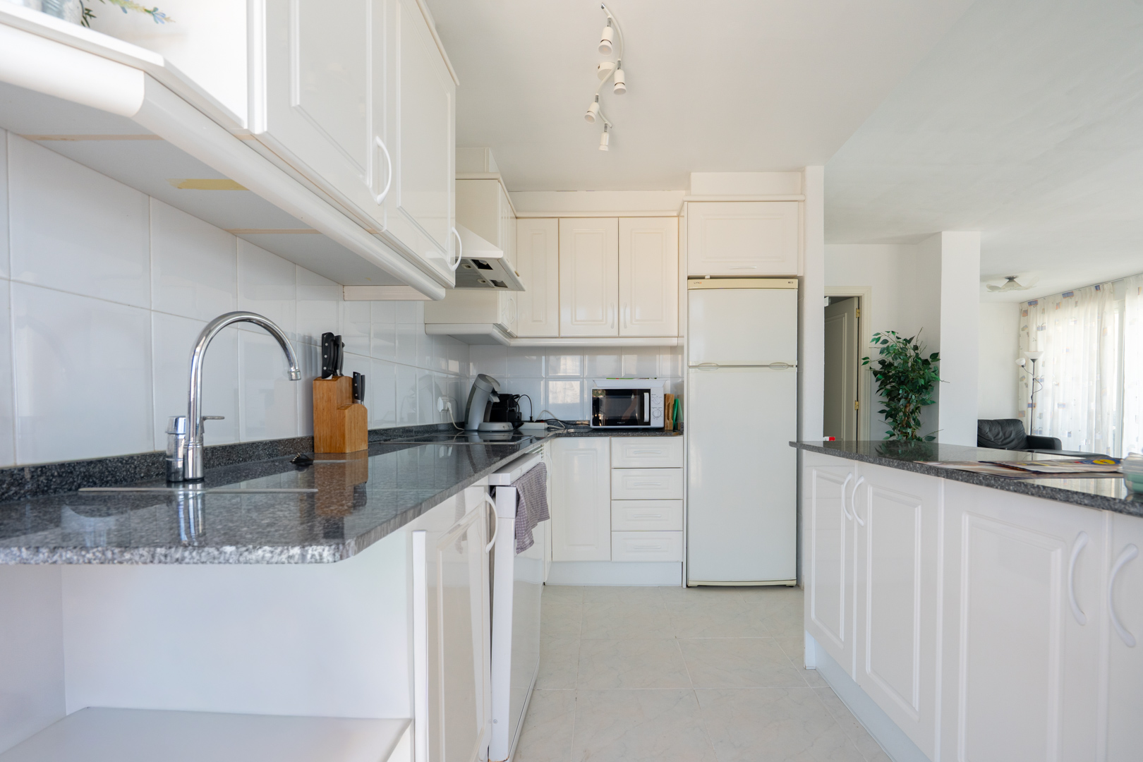 Cocina funcional con mobiliario blanco, encimeras de granito oscuro y azulejos blancos. Equipada con electrodomésticos esenciales y barra americana que conecta con el salón.